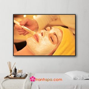 Mẫu tranh treo spa đẹp hình ảnh nhân viên đang đắp mặt nạ dưỡng da cho nữ khách hàng trong không gian với gam màu vàng sáng ấm áp sang trọng