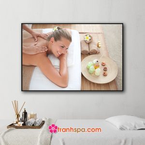Mẫu tranh treo spa đẹp hình ảnh nữ khách hàng đang được massage thư giãn lưng, phía trước là khay chứa tinh dầu và nến thơm, hoa sứ và những cuộn khăn tắm