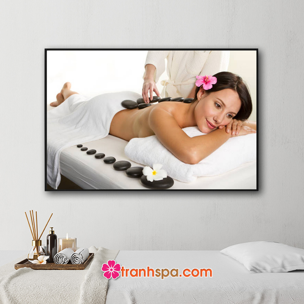 Mẫu tranh treo spa đẹp hình ảnh nhân viên đang chườm đá nóng trên lưng cho nữ khách hàng, bên cạnh giường là hàng đá cuội đen bóng và bông hoa sứ trắng