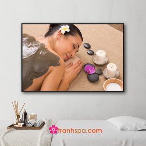Mẫu tranh treo spa đẹp hình ảnh nữ khách hàng đang được đắp bùn trên lưng để làm đẹp và thư giãn, xung quanh được trang trí với nến thơm, đá cuội, hoa lan tím và hoa sứ trắng cài trên tóc