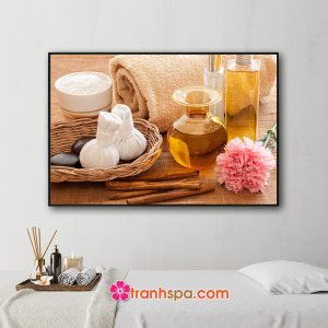 Mẫu tranh trang trí spa đẹp hình ảnh rổ mây chứa túi chườm và đá cuội, hoa cẩm chướng hồng, tinh dầu, muối tắm, thanh quế và khăn tắm cuộn tròn