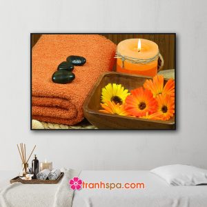 Mẫu tranh trang trí spa đẹp hình ảnh những viên đá cuội đặt trên khăn tắm, nến thơm và chậu nước rải những cánh hoa cúc màu vàng và cam