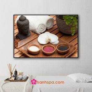 Mẫu tranh trang trí spa đẹp hình ảnh đầu tượng phật, chậu lá xanh, khăn tắm cuộn, khay đựng hoa lan và các loại muối tắm đặt trên nền gỗ