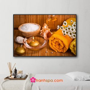 Mẫu tranh trang trí spa đẹp hình ảnh bát muối tắm, tinh dầu, đá cuội, vỏ ốc biển, cuộn khăn tắm, hoa cúc trắng, nến thơm tỏa ánh sáng vàng ấm áp trên nền mành trúc